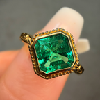 Gold ring with a green gemstone held between fingers against a neutral background