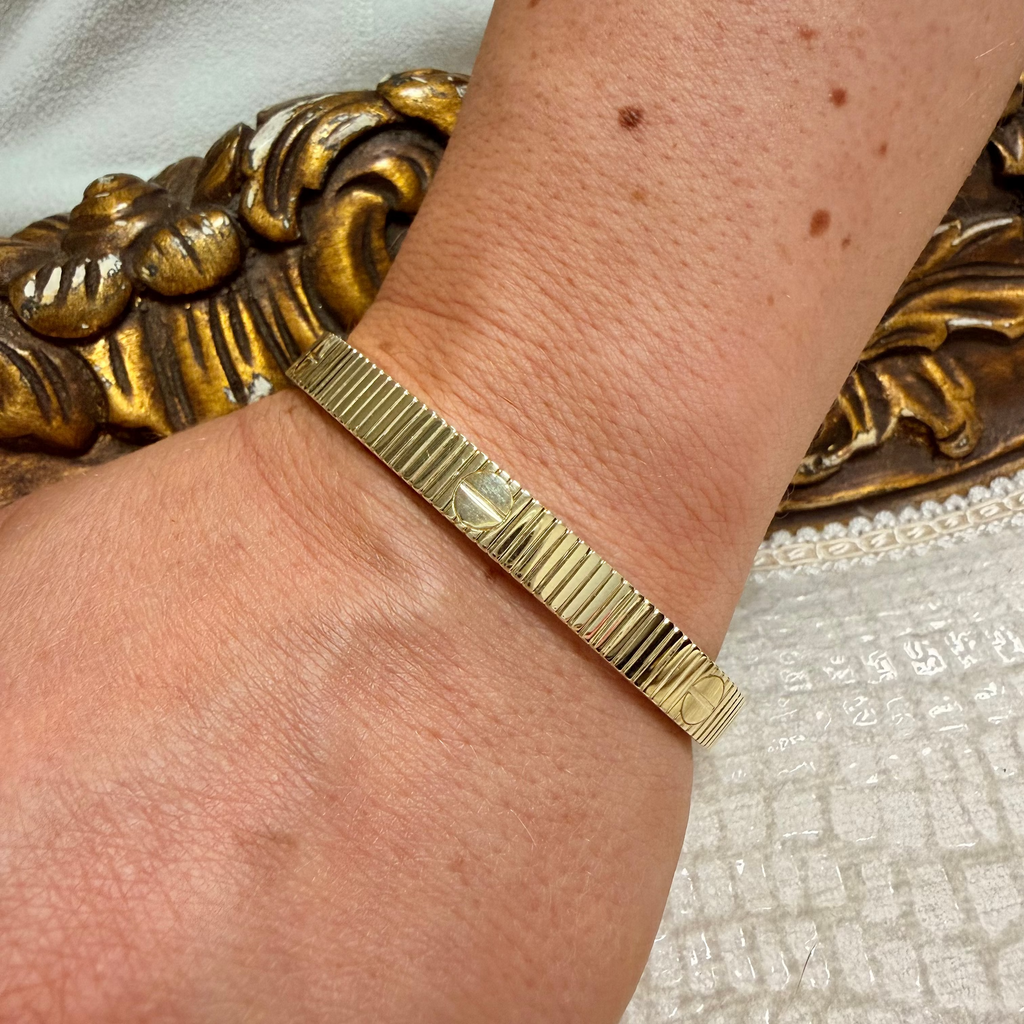 A close-up of a person's wrist wearing a textured gold bangle bracelet, with a white textured fabric and an ornate gold decorative surface in the background.