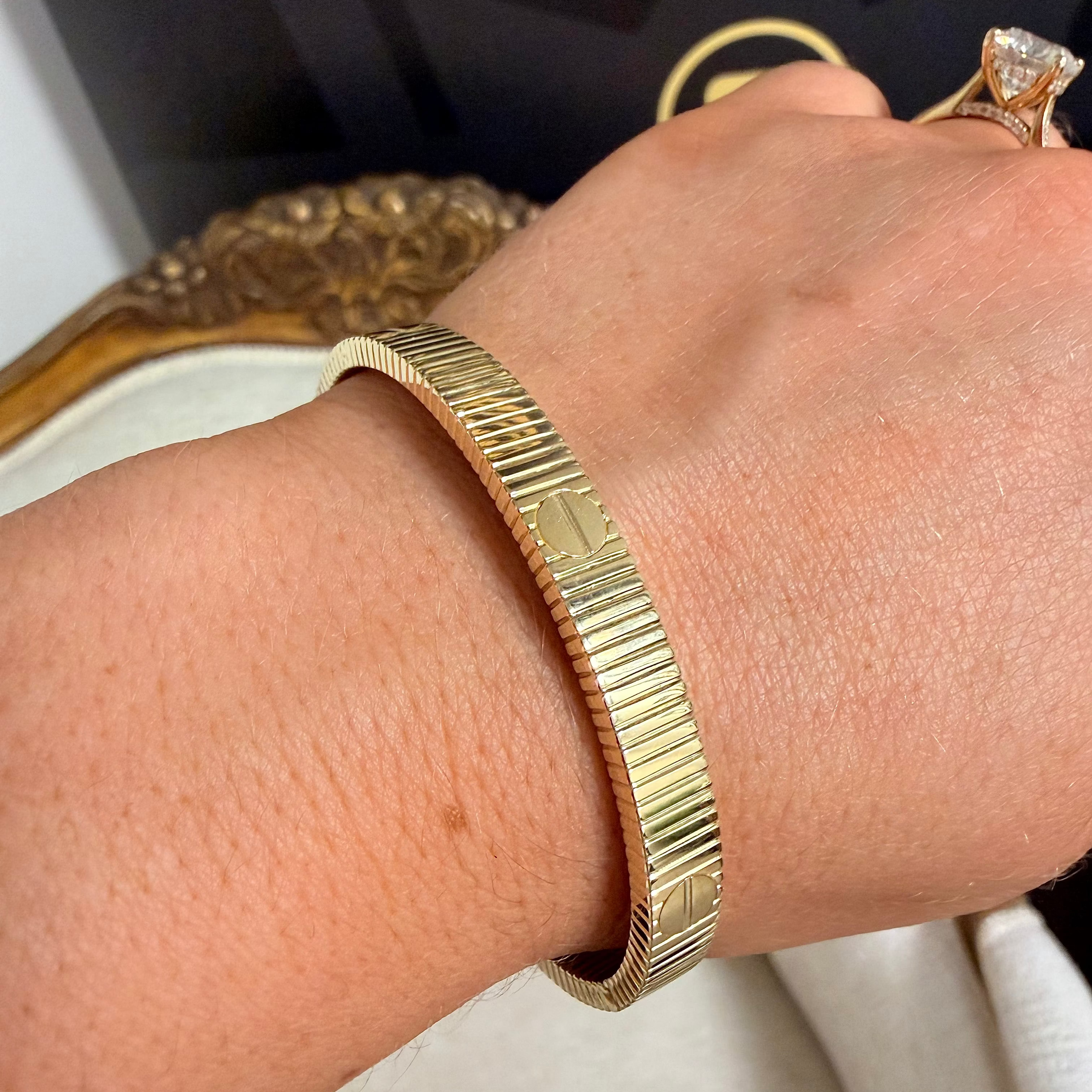 A gold-plated mobile jewelry bangle bracelet with a fluted, ridged texture, shown worn on a person's wrist. A gold ring is also visible on the finger, with a black jewelry box in the background.
