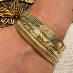Three gold-toned textured bangles stacked on a person's wrist, with a close-up of the skin and part of a decorative gold object in the background.
