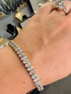 A close-up of a person's wrist wearing a diamond tennis bracelet, with a gold ring on their finger. The background shows a blurred jewelry display.