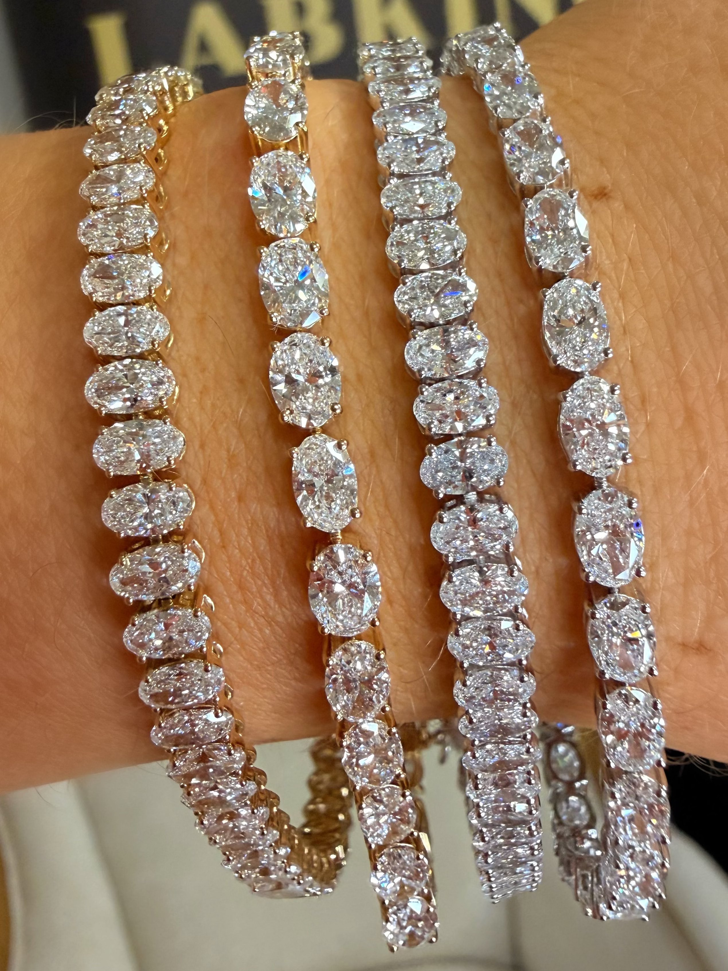 Four diamond tennis bracelets in different metal settings (yellow gold, rose gold, white gold, and silver) worn stacked on a person's wrist.
