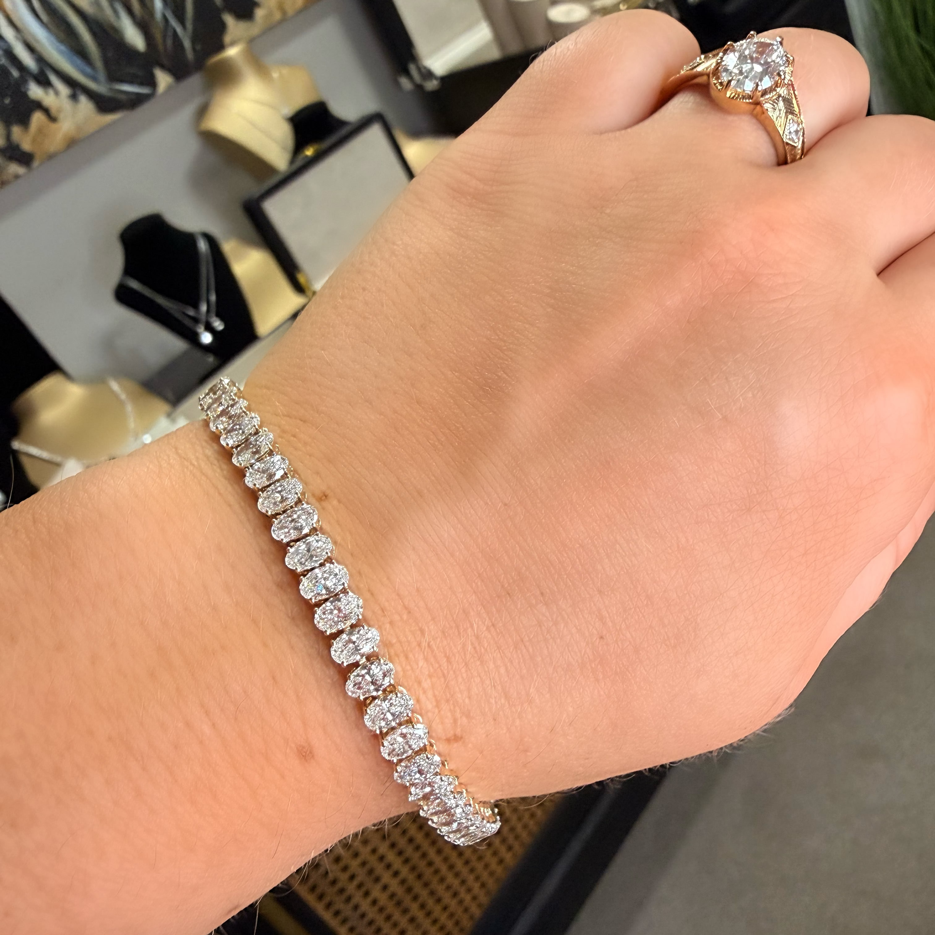 A close-up of a person's hand wearing a sparkling diamond tennis bracelet, with a gold ring on their finger. The background shows a jewelry display with mannequins and boxes.