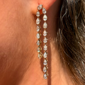 A close-up of a person's ear wearing a pair of long gold drop earrings with a series of clear teardrop-shaped stones.
