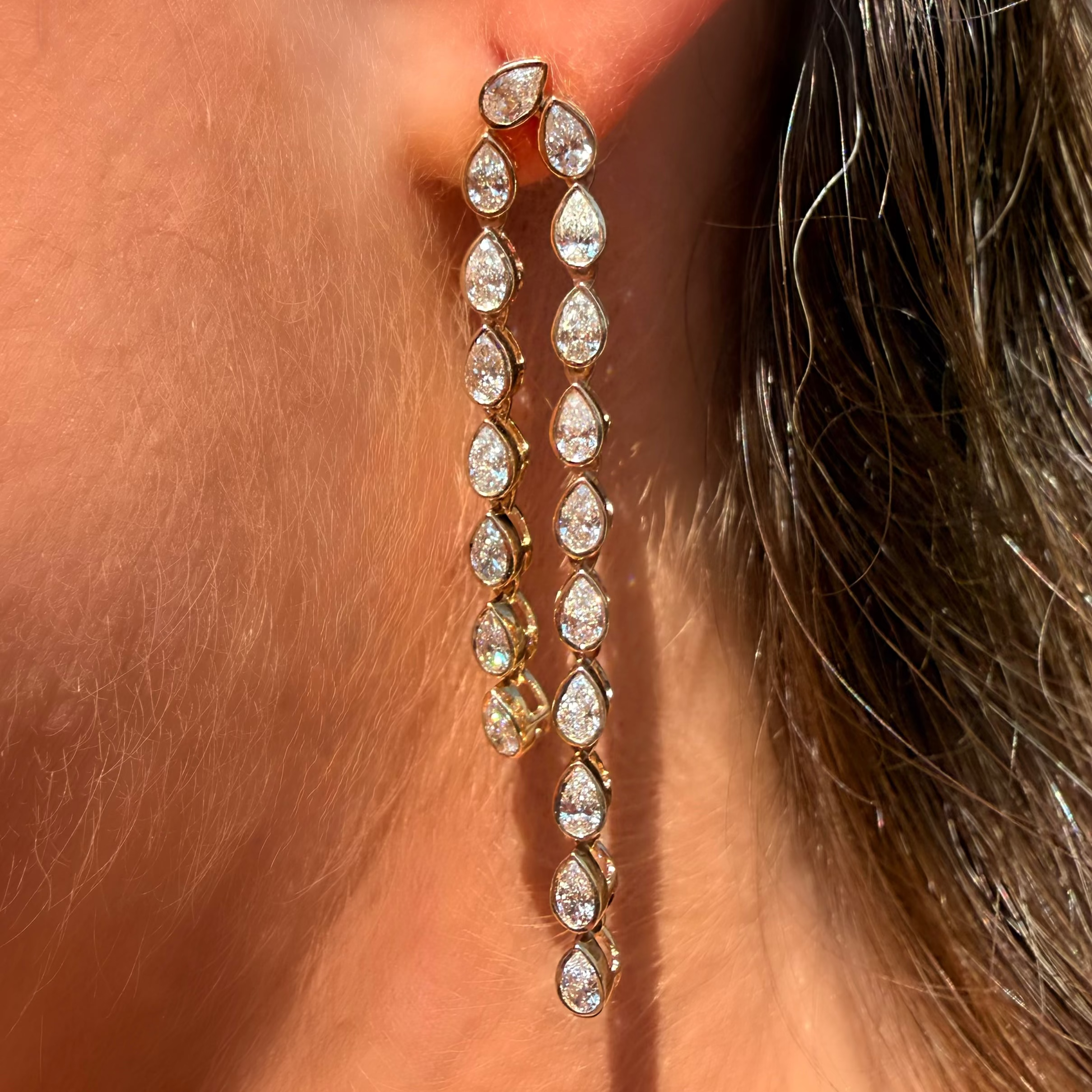 A close-up of a person's ear wearing a pair of long gold drop earrings with a series of clear teardrop-shaped stones.