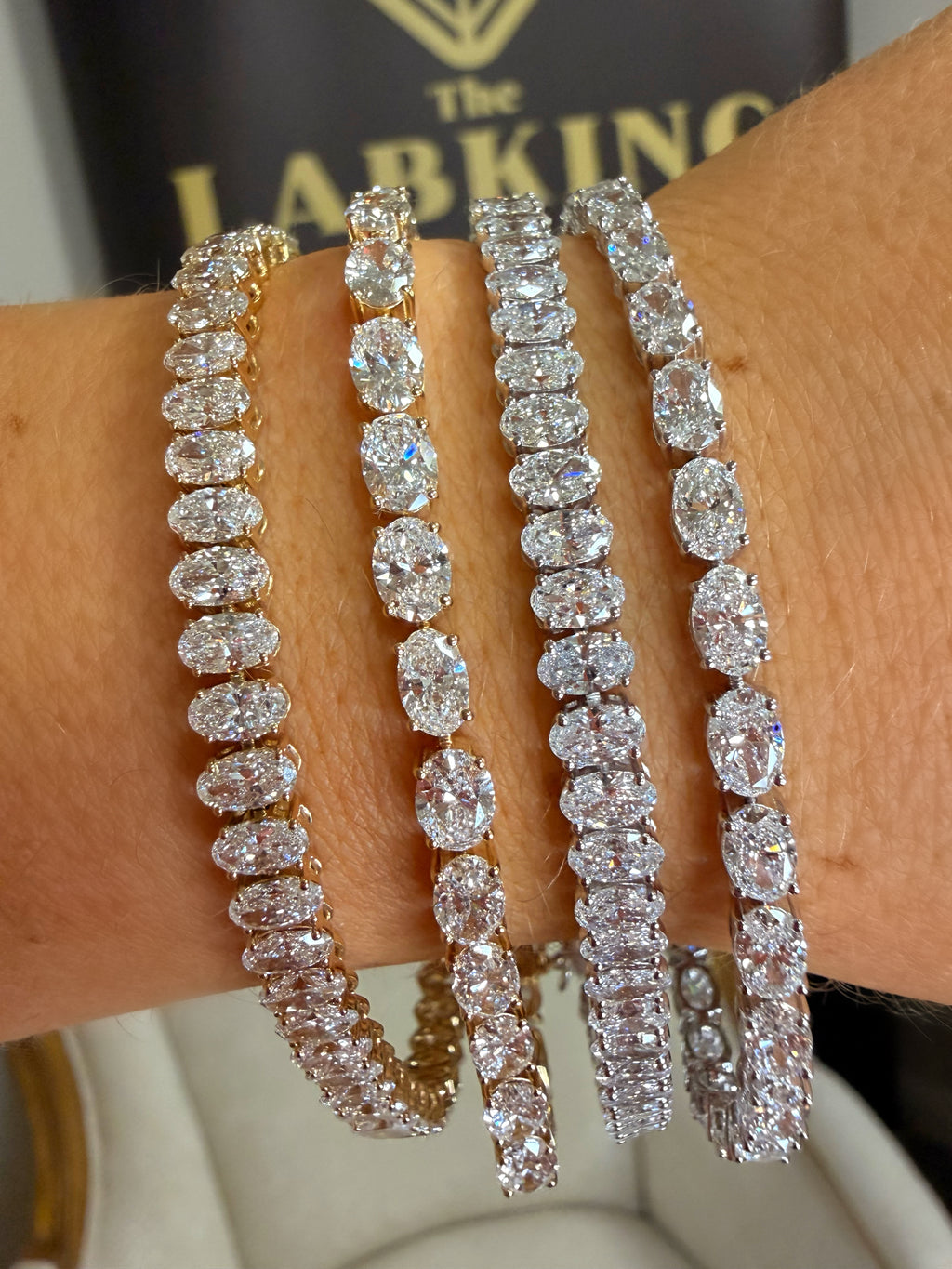 Four diamond tennis bracelets in different metal settings (yellow gold, rose gold, white gold, and silver) worn stacked on a person's wrist.