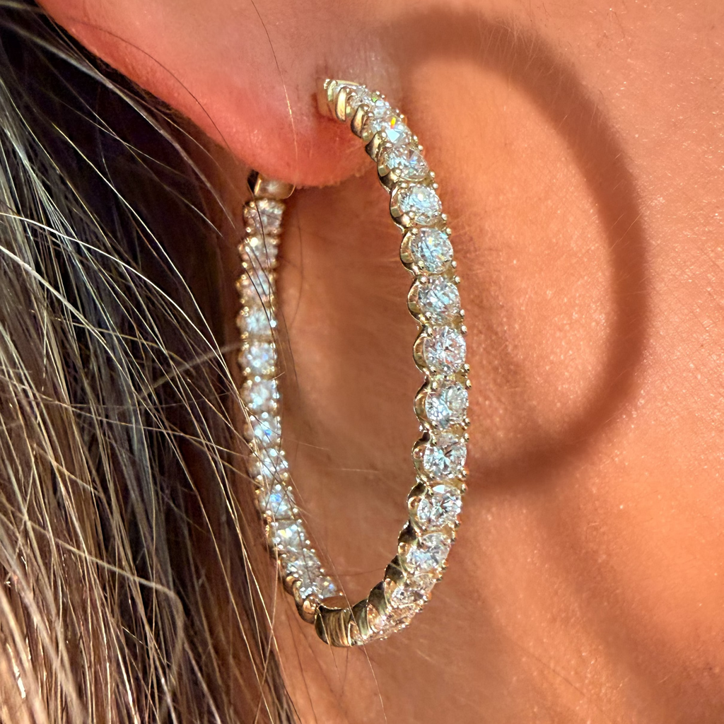 A close-up of a person's ear wearing a large gold hoop earring set with clear pavé gemstones. The earring is shown against warm skin tones and dark hair.