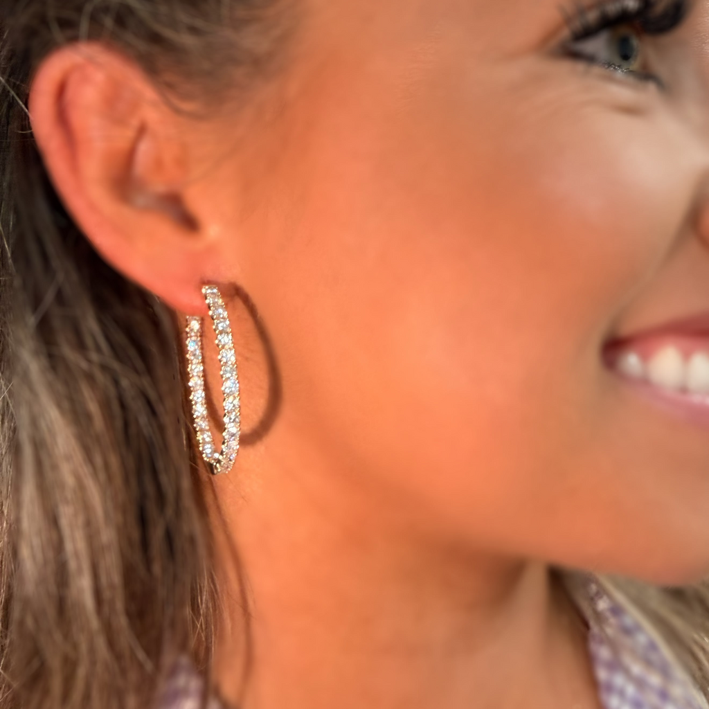 A close-up side profile of a woman wearing a pair of silver-toned hoop earrings adorned with clear crystals, set against a warm, sunlit background.