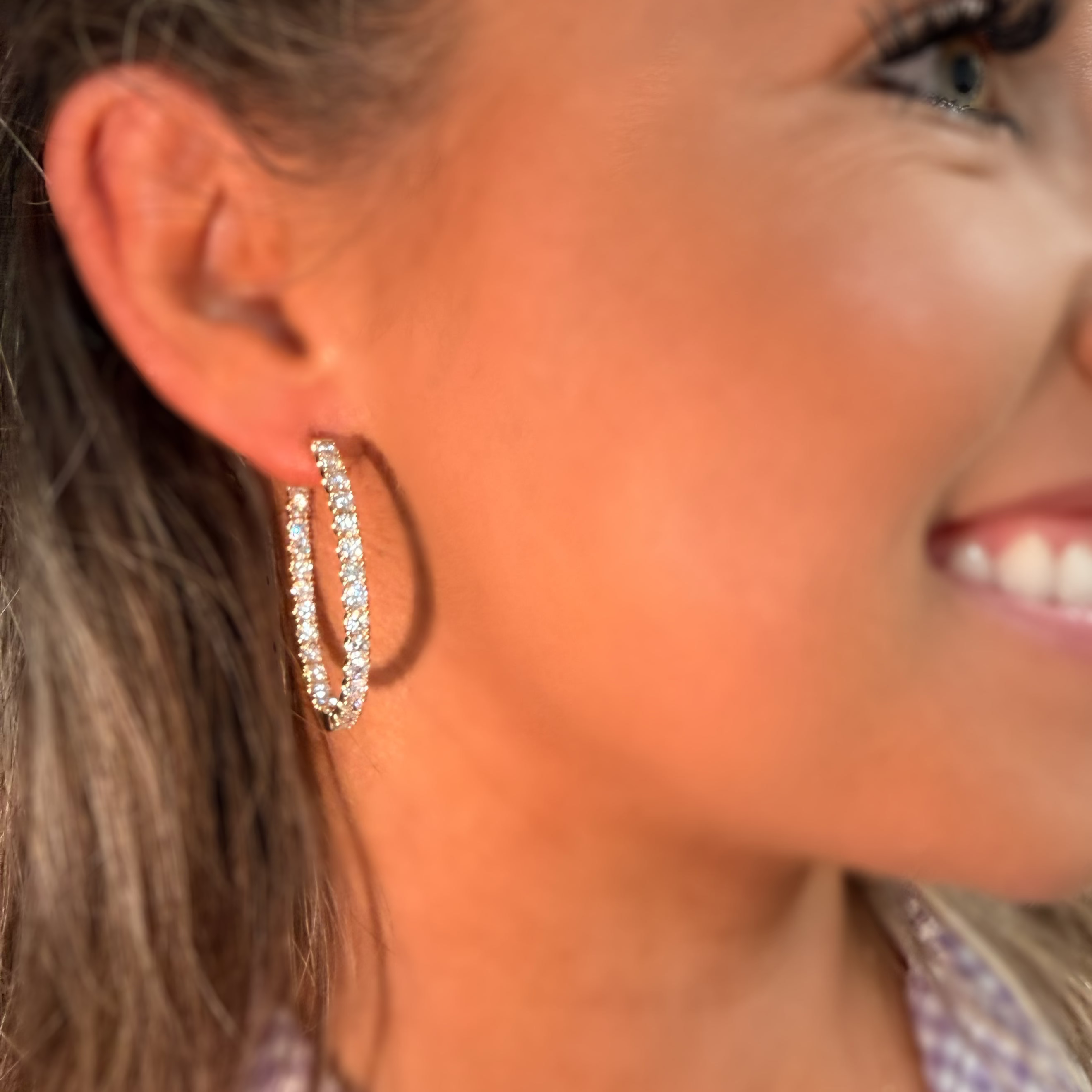 A close-up side profile of a woman wearing a pair of silver-toned hoop earrings adorned with clear crystals, set against a warm, sunlit background.