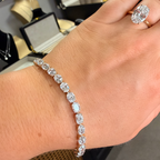 A close-up of a person's hand wearing a diamond tennis bracelet on the wrist, with a matching diamond ring on the index finger. The jewelry is set in yellow gold.