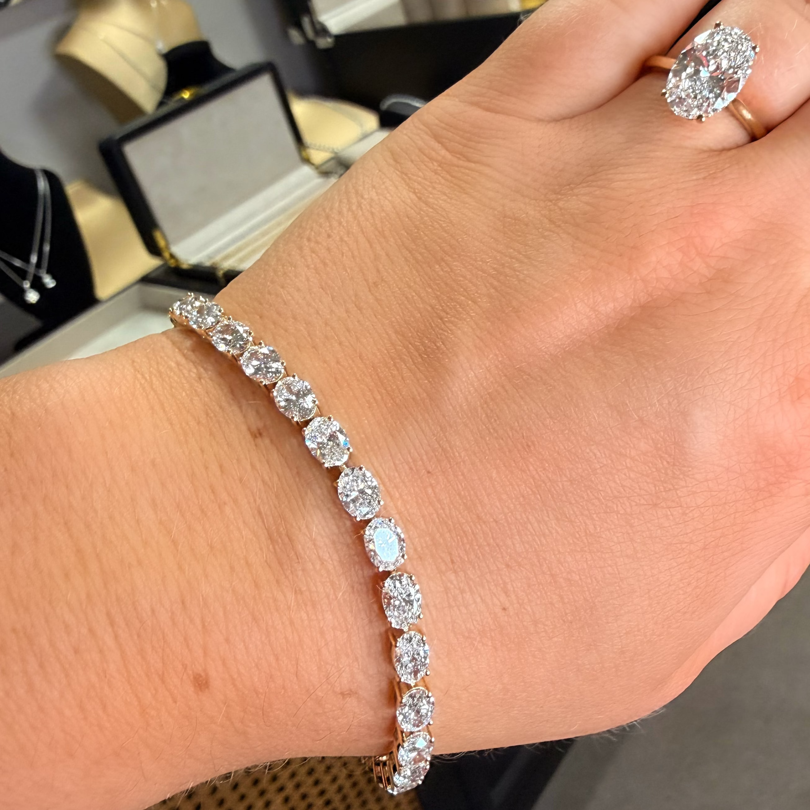 A close-up of a person's hand wearing a diamond tennis bracelet on the wrist, with a matching diamond ring on the index finger. The jewelry is set in yellow gold.