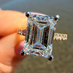 A close-up of a hand holding a large emerald-cut diamond ring with a silver-toned band and small side diamonds, with a blurred outdoor background.