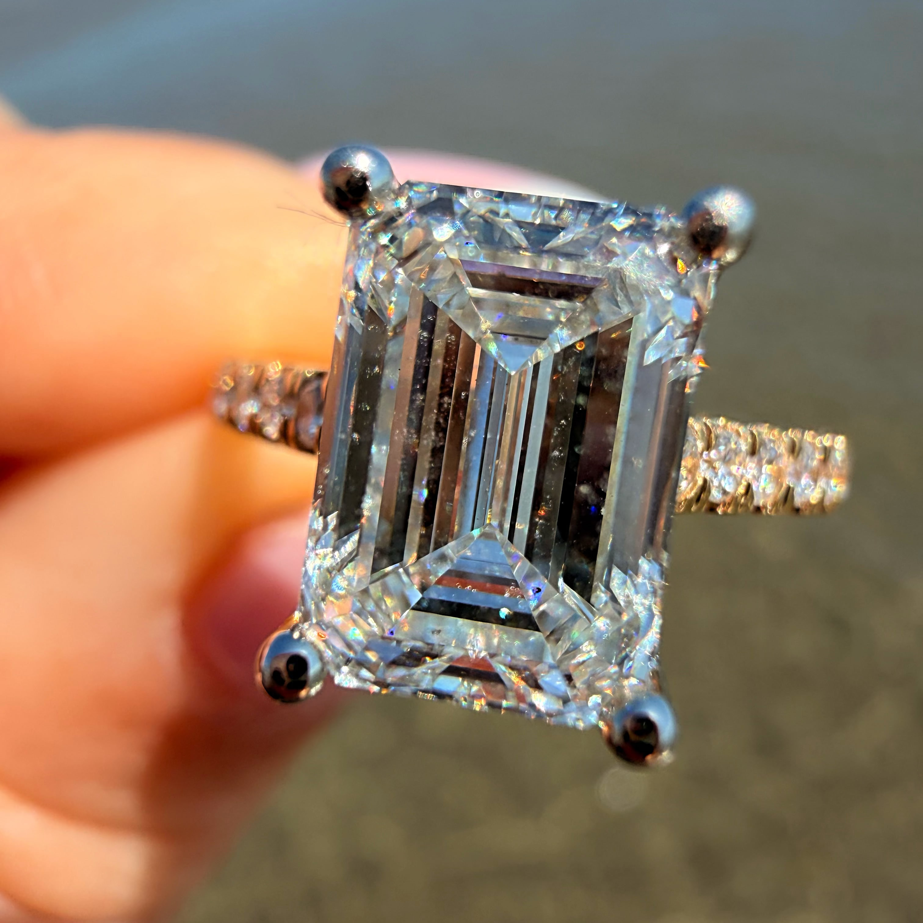 A close-up of a hand holding a large emerald-cut diamond ring with a silver-toned band and small side diamonds, with a blurred outdoor background.