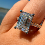 An emerald-cut diamond ring with a silver-toned band, worn on a finger outdoors with a blue sky and water in the background.