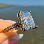 A close-up of a hand holding a rose gold ring with a large emerald-cut diamond center stone and smaller diamonds along the band, set against a blurred outdoor background of water and sky.