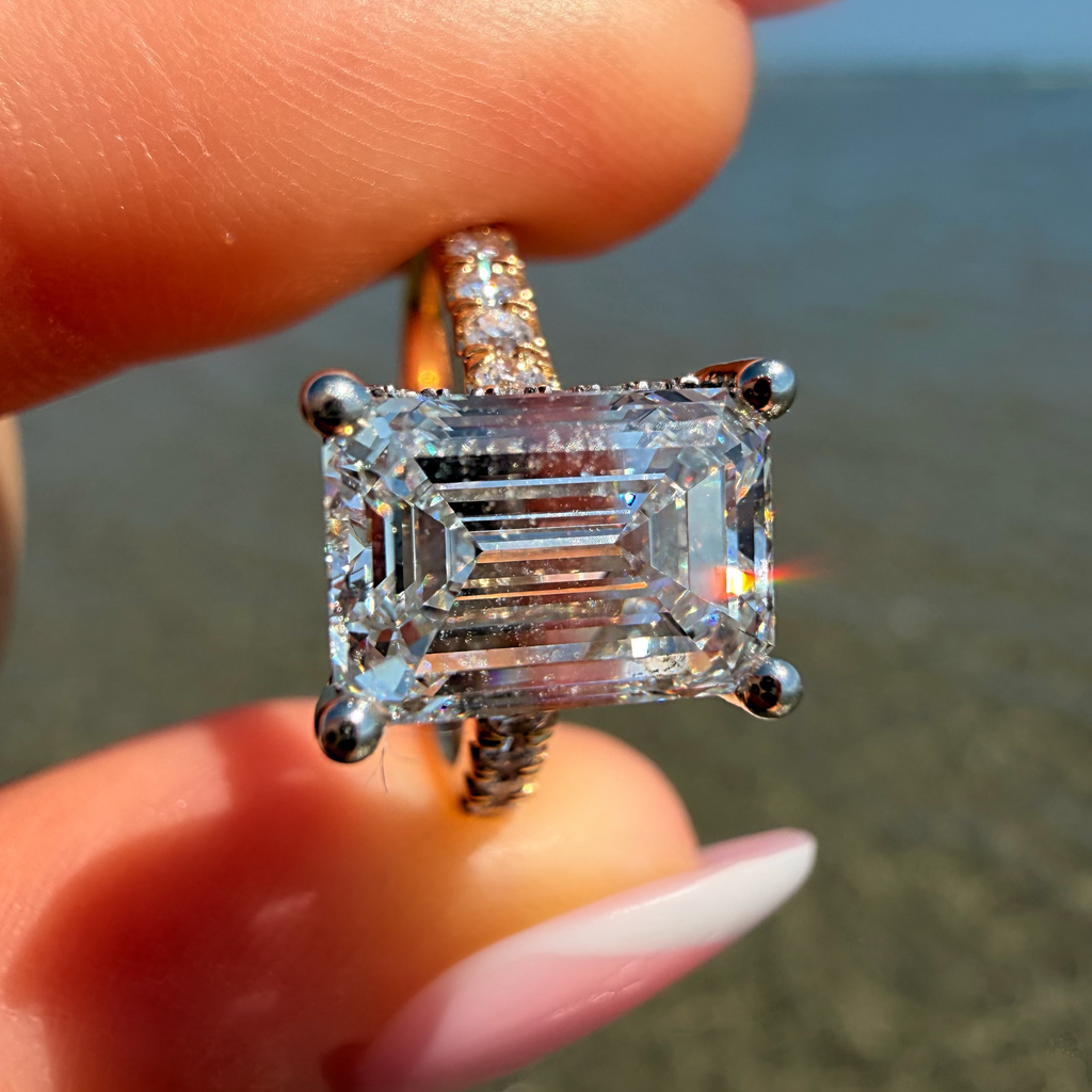 A close-up of a hand holding a ring with a large rectangular emerald-cut clear gemstone in a silver-toned setting, with a beach and blue sky blurred in the background.