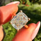 A close-up of a cushion-cut diamond ring held between fingers outdoors. The ring features a brilliant clear gemstone set in a yellow gold four-prong setting, with a blurred green garden background.