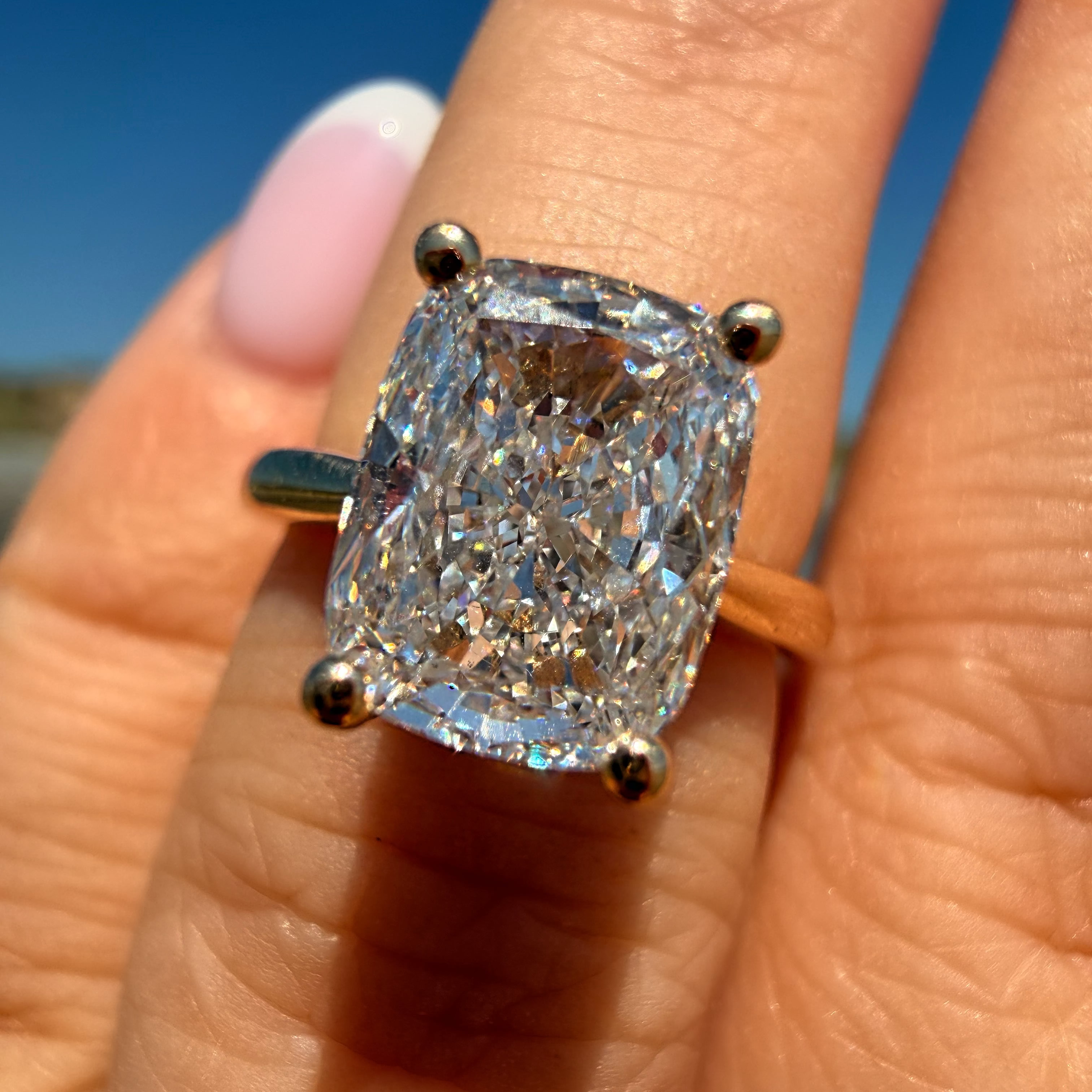 A close-up of a hand wearing a large cushion-cut diamond ring with a yellow gold band, set against a bright blue sky background.