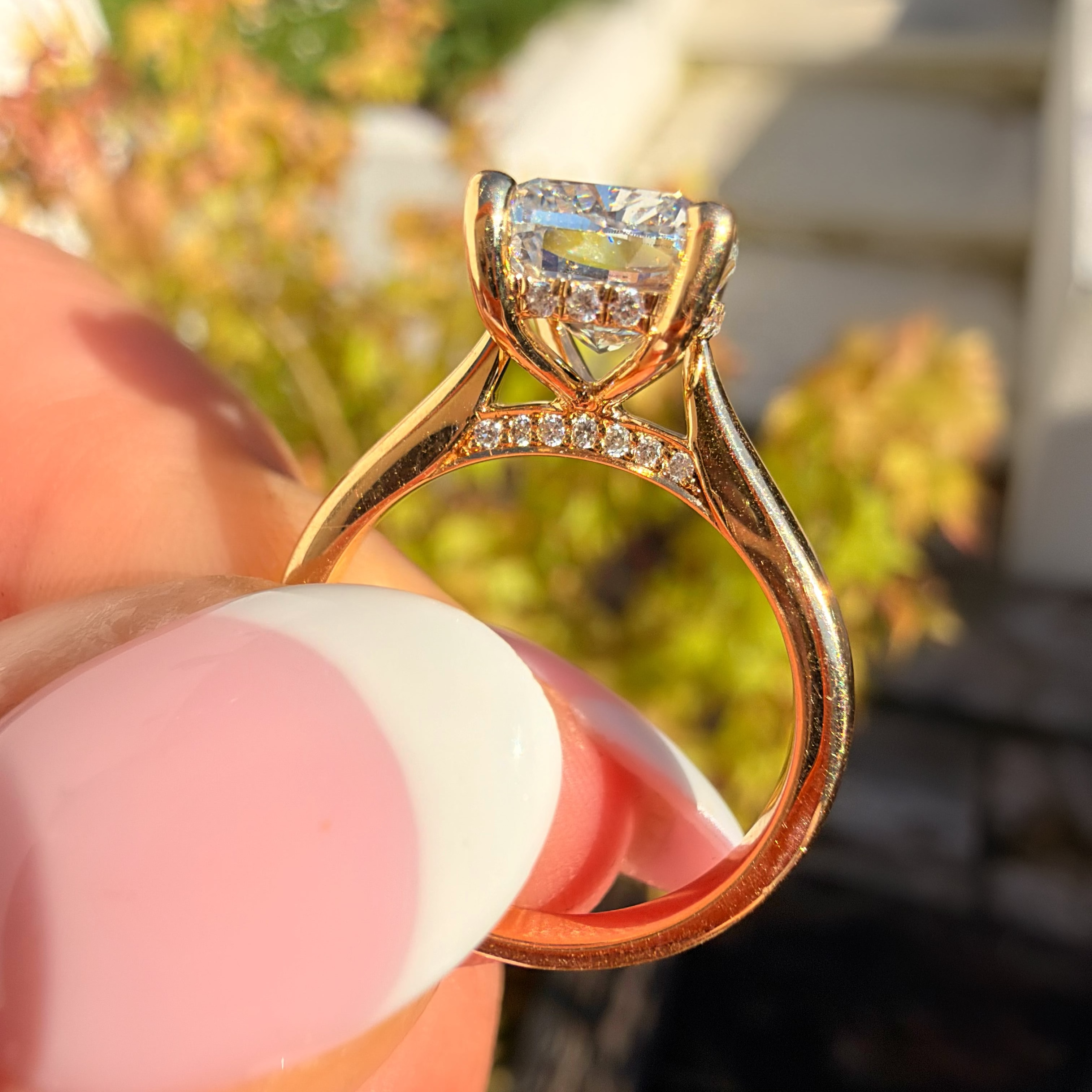 A close-up shot of a gold ring with a clear heart-shaped gemstone held by a hand with pink and white painted fingernails, set against a blurred outdoor background of green foliage and light-colored steps.