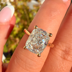 A close-up of a hand wearing a yellow gold ring with a cushion-cut clear gemstone, set against a blurred outdoor background.