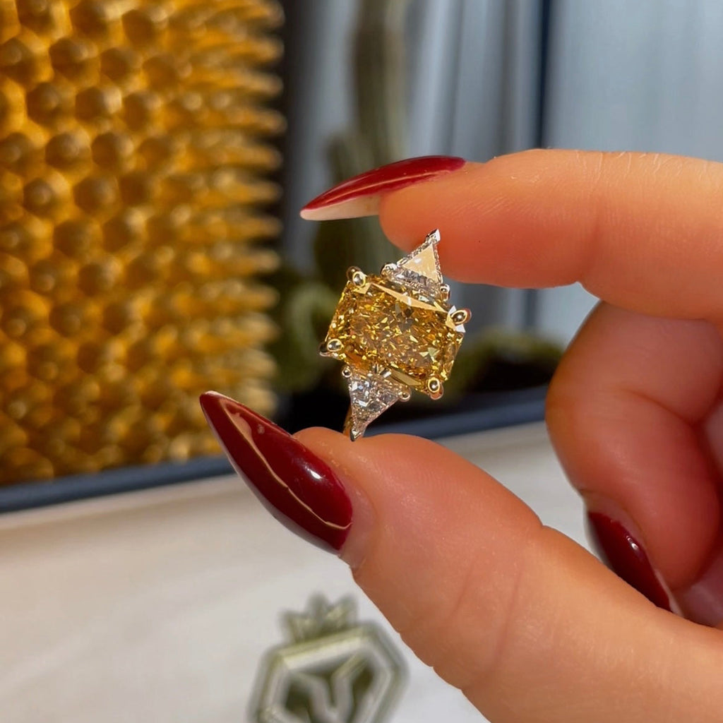 Gold and diamond ring held by a hand with red nail polish, brand logo visible in the background.