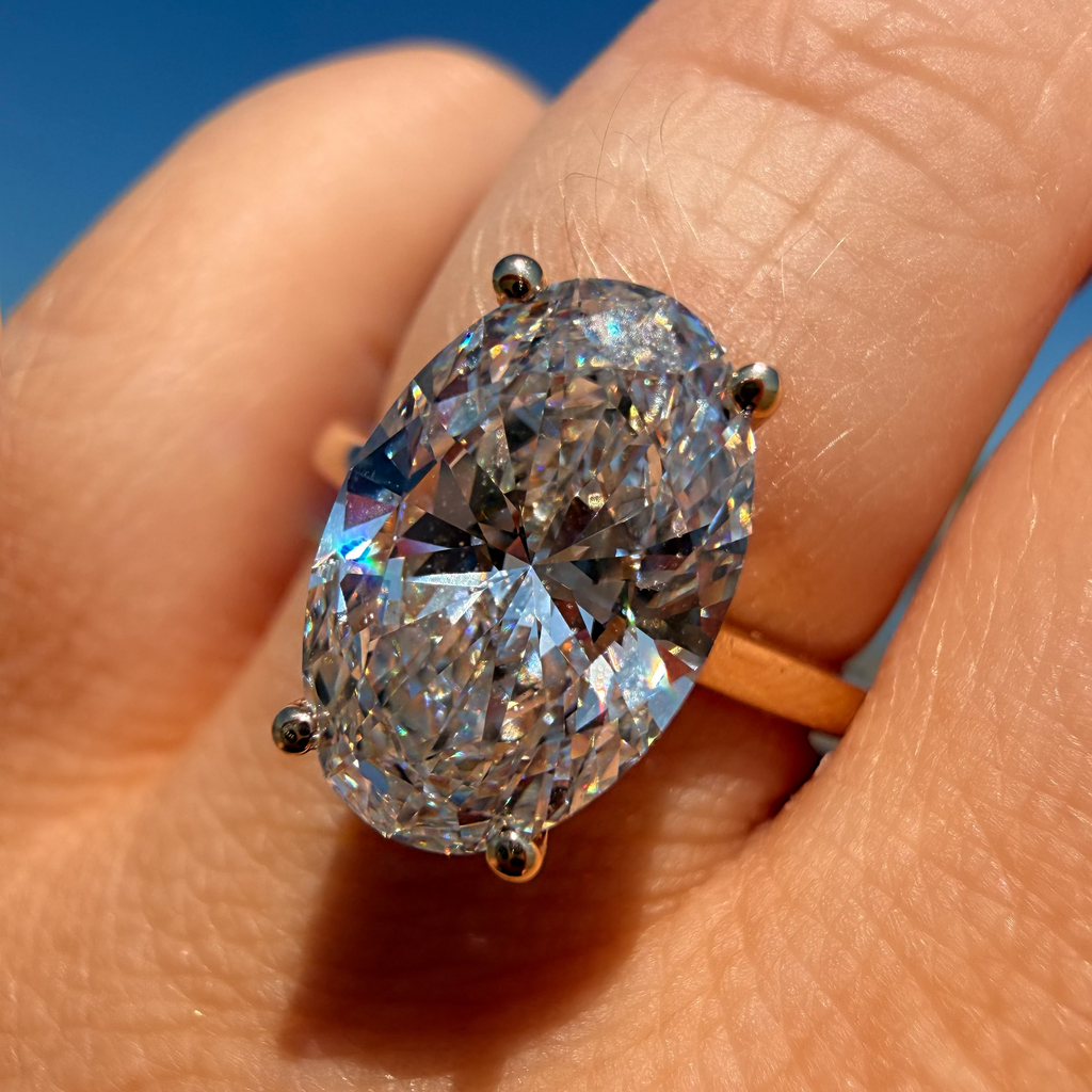 An oval-cut clear gemstone ring with a yellow gold band, worn on a person's finger against a bright blue sky background.