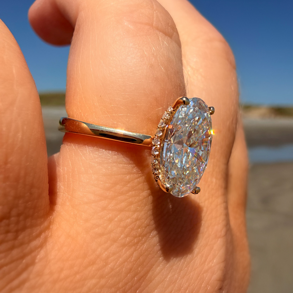 An oval-cut diamond ring with a yellow gold band and a halo of small diamonds, shown worn on a person's hand against a beach and blue sky background.