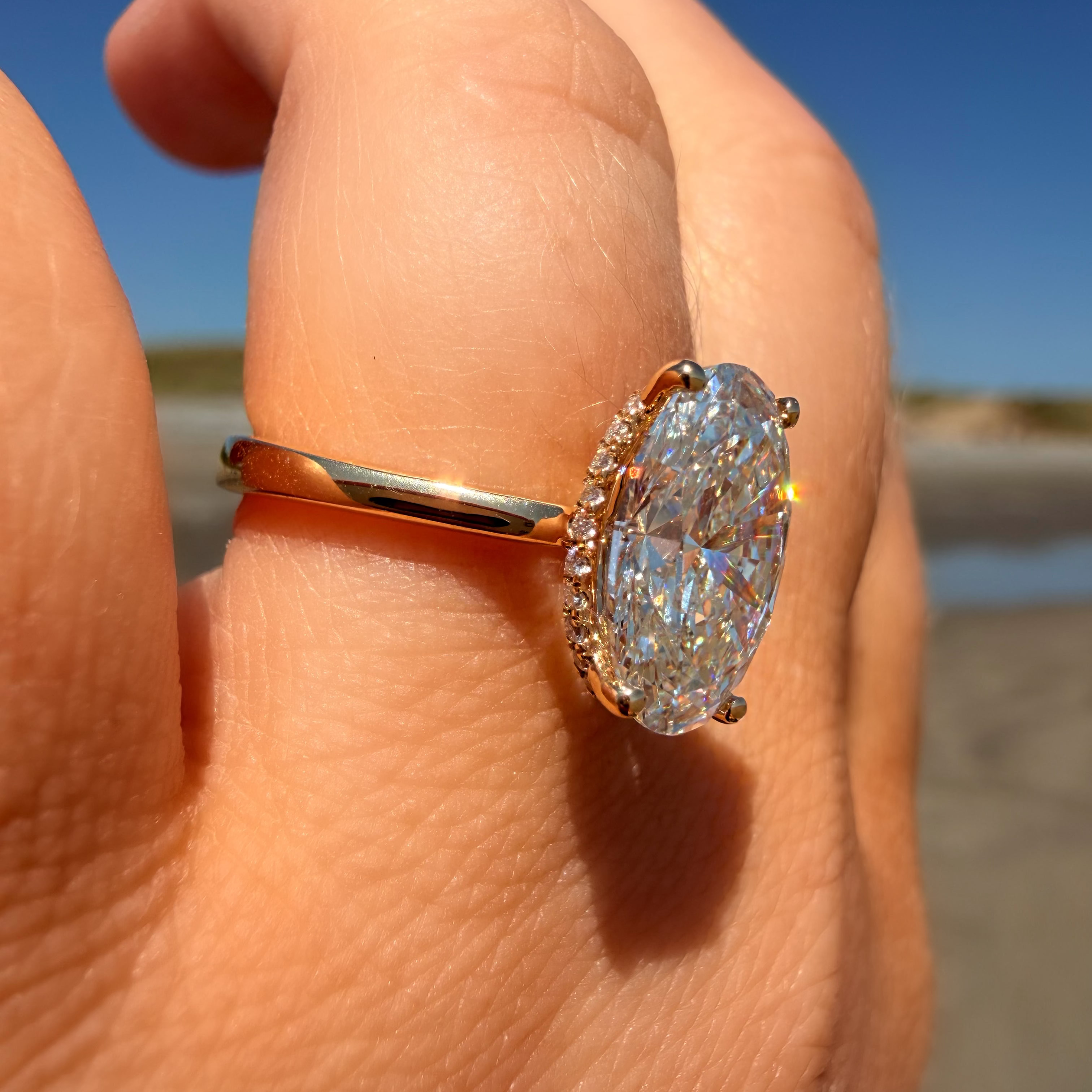 An oval-cut diamond ring with a yellow gold band and a halo of small diamonds, shown worn on a person's hand against a beach and blue sky background.