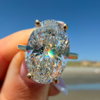 A close-up of a hand holding a large oval-cut diamond ring with a silver-toned band, set against a blurred beach and blue sky background.