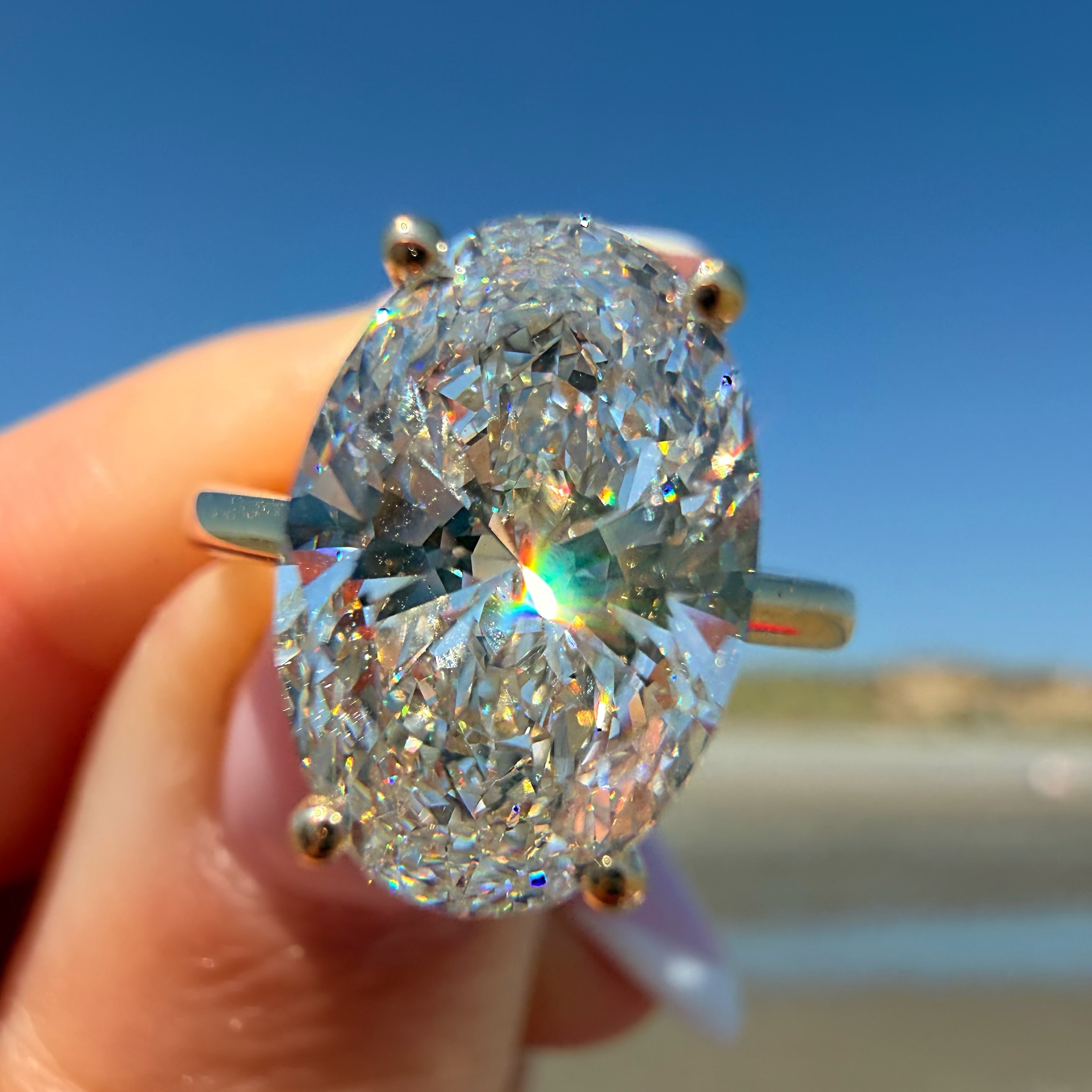 A close-up of a hand holding a large oval-cut diamond ring with a silver-toned band, set against a blurred beach and blue sky background.