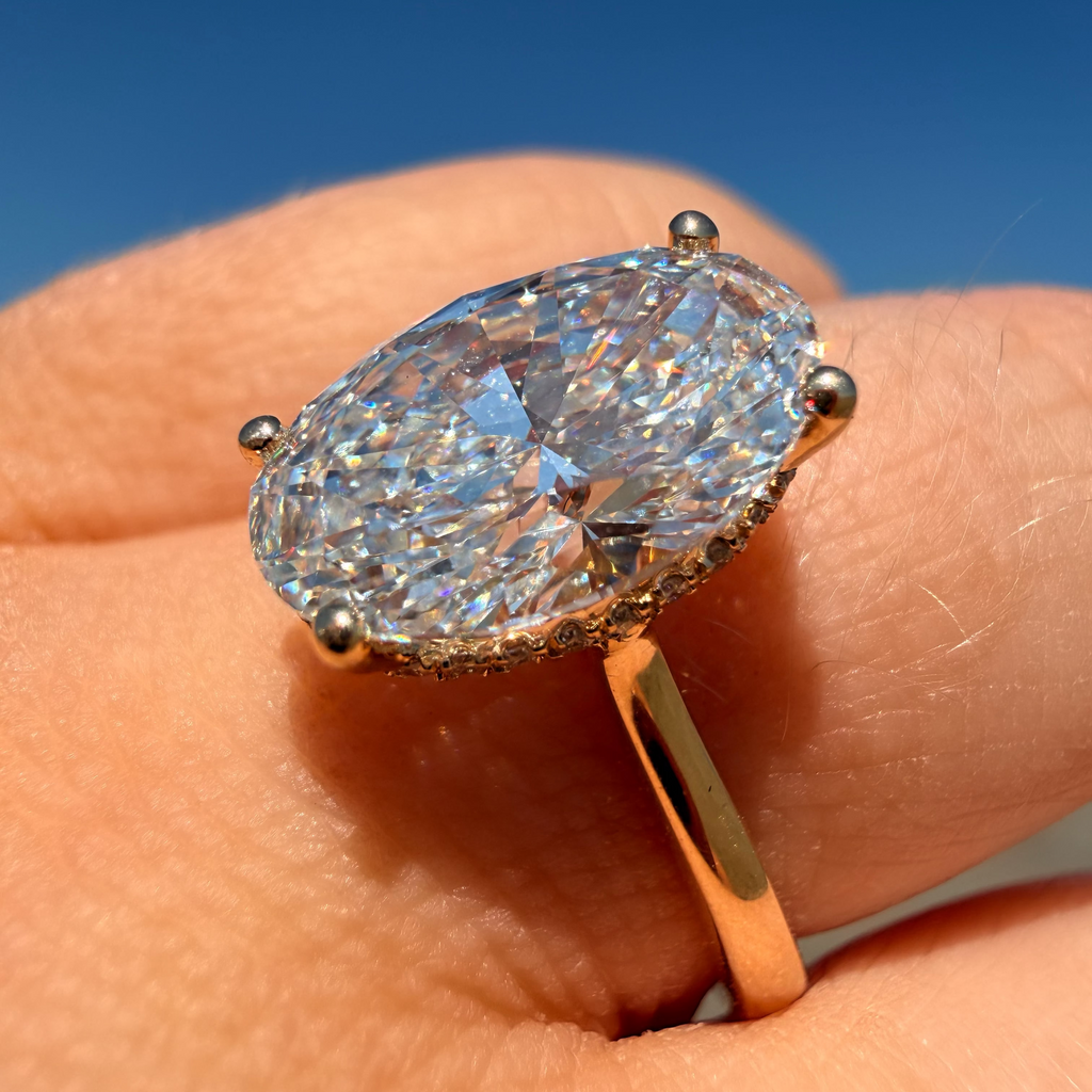 An oval-cut diamond ring with a yellow gold band, worn on a finger against a clear blue sky background.