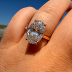 A close-up of a hand wearing a large oval diamond ring with a yellow gold band, set against a bright blue sky background.
