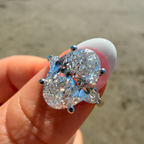 A close-up of a hand holding a silver ring with a large oval-cut clear gemstone and smaller marquise-cut side stones, with a blurred beach background.