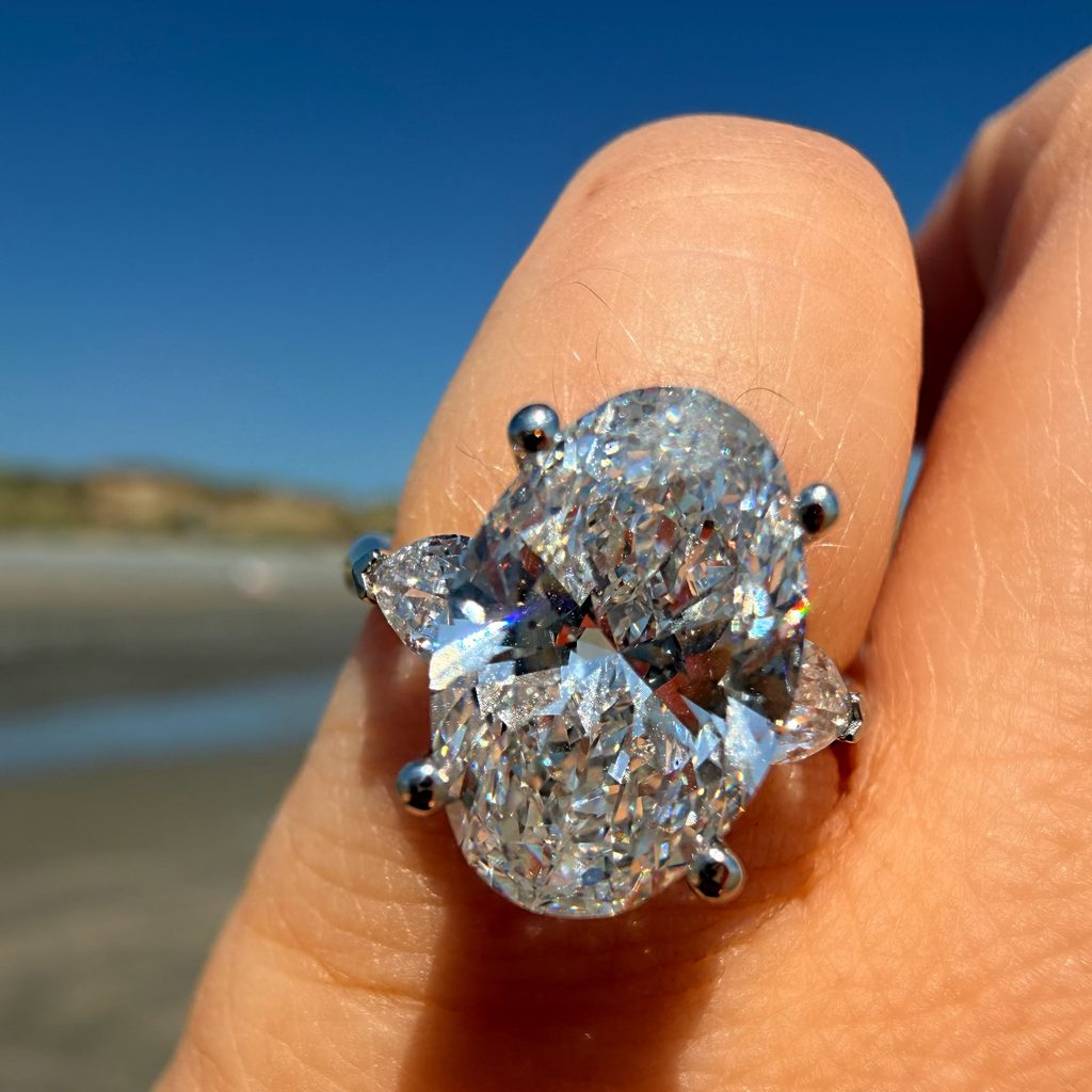 A close-up of a hand wearing a large oval diamond ring with pear-shaped side stones in a silver-toned setting, photographed outdoors against a blurred beach and blue sky background.