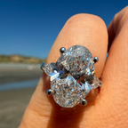 A close-up of a hand wearing a large oval diamond ring with pear-shaped side stones in a silver-toned setting, photographed outdoors against a blurred beach and blue sky background.