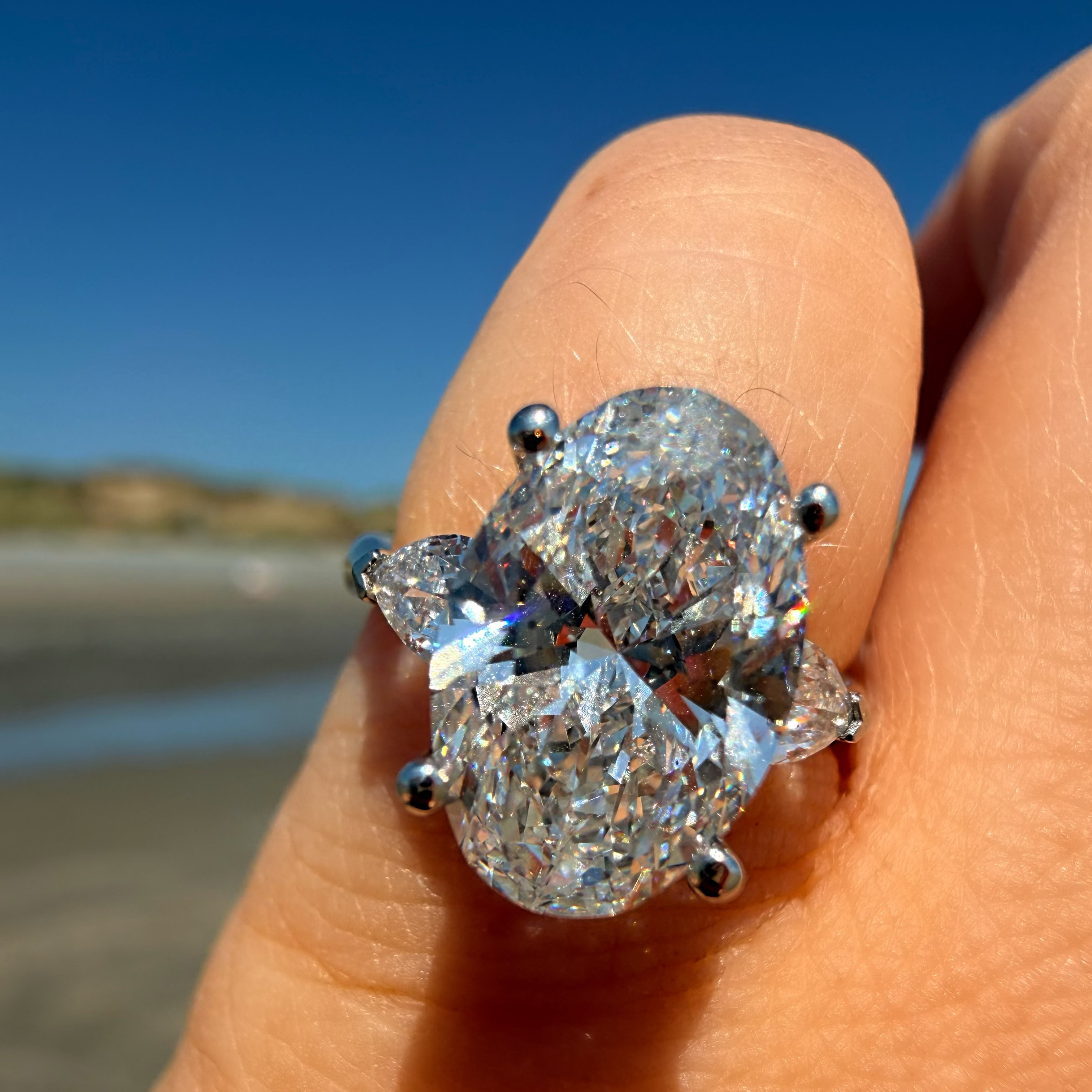 A close-up of a hand wearing a large oval diamond ring with pear-shaped side stones in a silver-toned setting, photographed outdoors against a blurred beach and blue sky background.