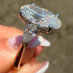 A close-up of a hand holding a diamond ring with a large oval center stone and a smaller marquise-cut side stone in a silver-toned setting, against a blurred outdoor background.
