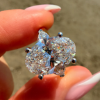 A close-up of a hand holding a brilliant round-cut diamond ring with a silver-toned prong setting against a blurred outdoor background.