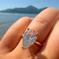 A pear-cut diamond ring with a gold band worn on a hand, with a blurred beach and mountain background.