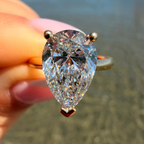 A close-up of a hand holding a pear-cut diamond ring with a rose gold setting against a blurred beach background.