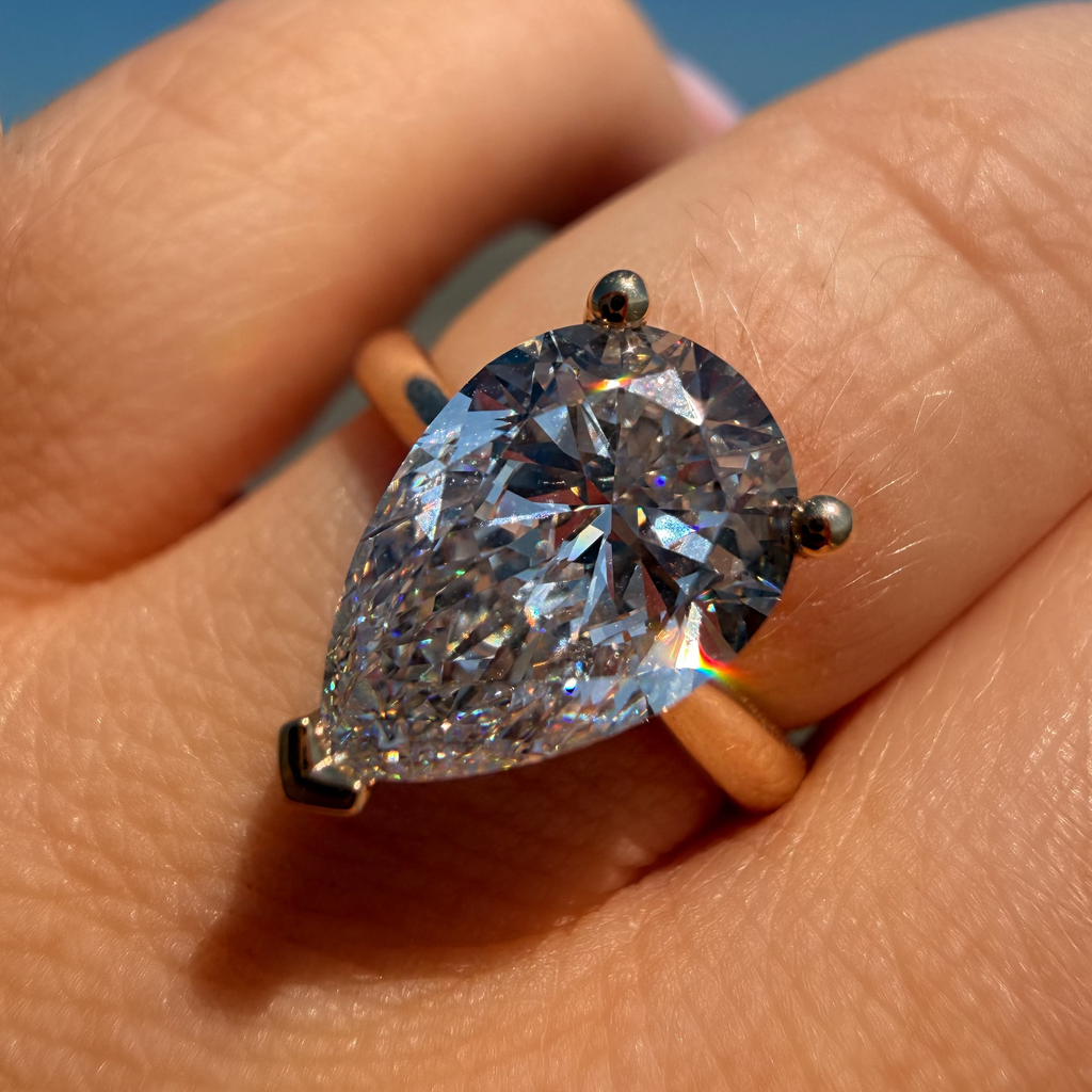 A pear-cut diamond ring in a yellow gold setting, worn on a person's finger against a blue sky background.