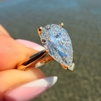 A close-up of a hand holding a pear-cut blue gemstone ring in a rose gold setting, with a blurred beach background.