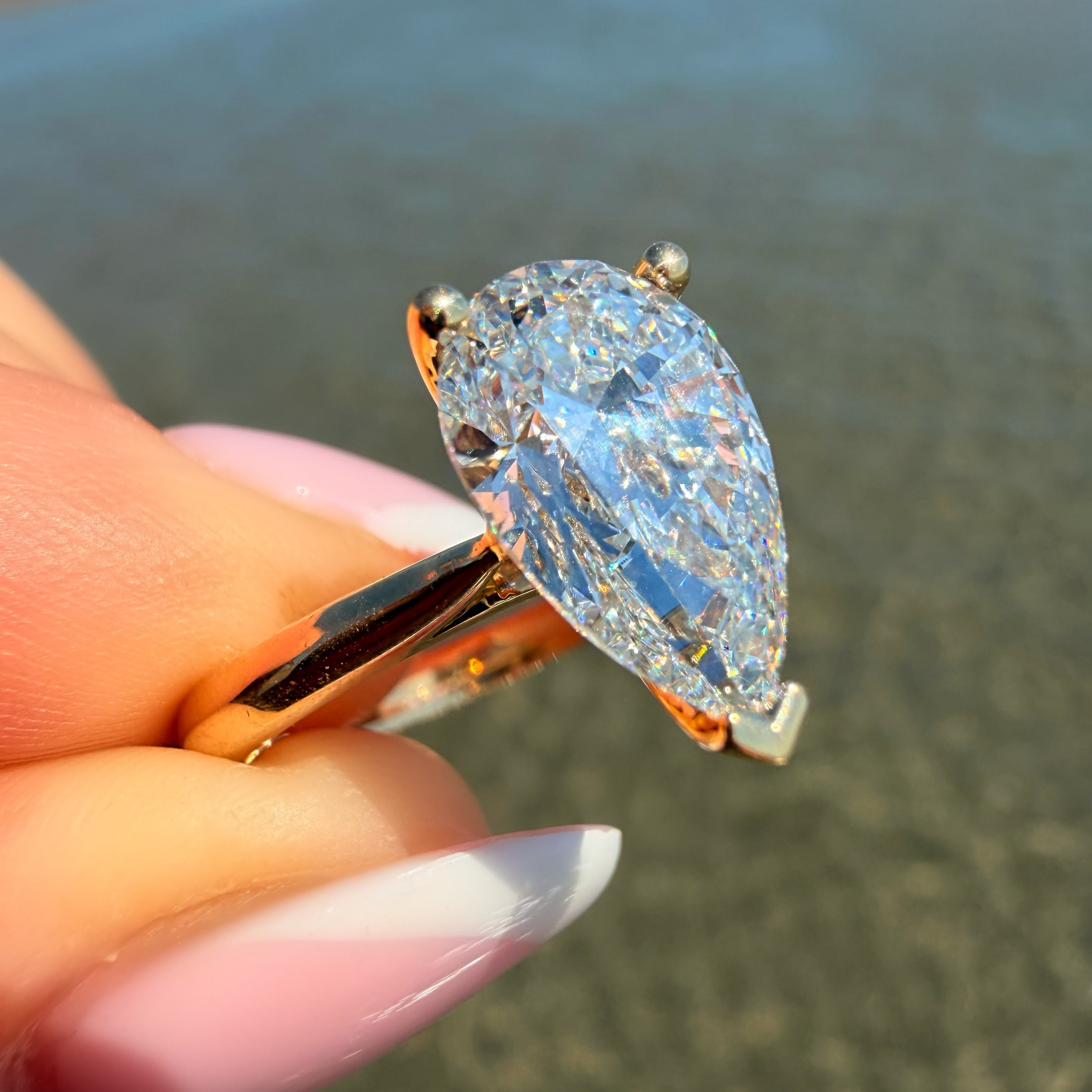 A close-up of a hand holding a pear-cut blue gemstone ring in a rose gold setting, with a blurred beach background.
