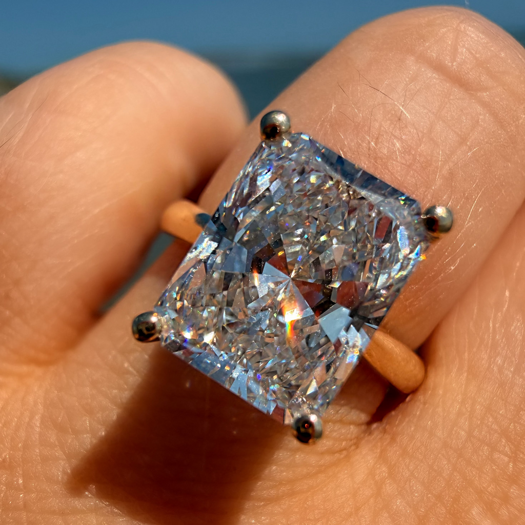 A close-up of a hand wearing a ring with a large rectangular clear gemstone in a yellow metal setting, photographed outdoors against a blurred blue sky background.