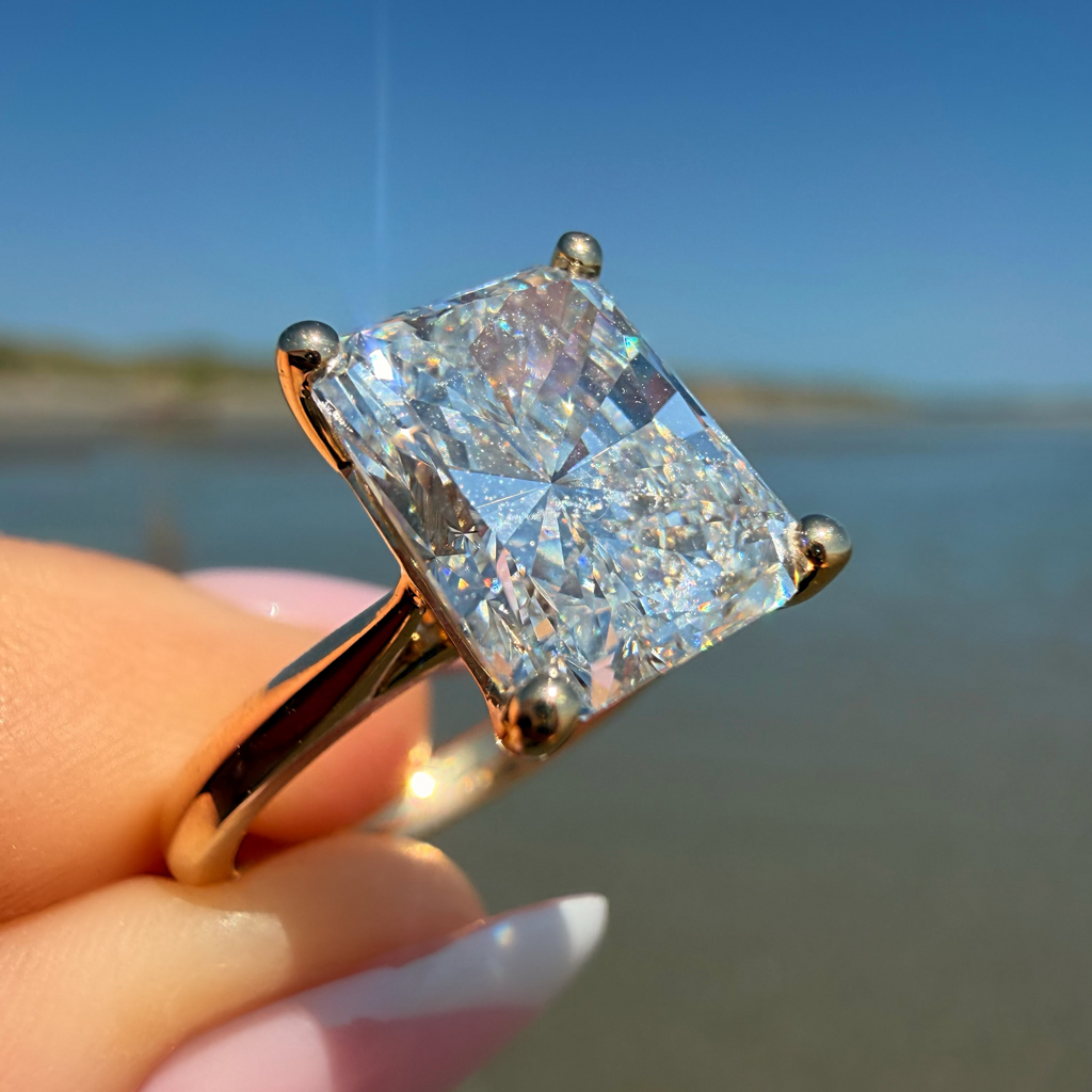 A gold ring with a large square clear gemstone held by a hand against a blurred beach and blue sky background.