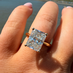 A close-up of a hand wearing a yellow gold ring with a large, brilliant-cut clear gemstone, set against a blurred beach background.