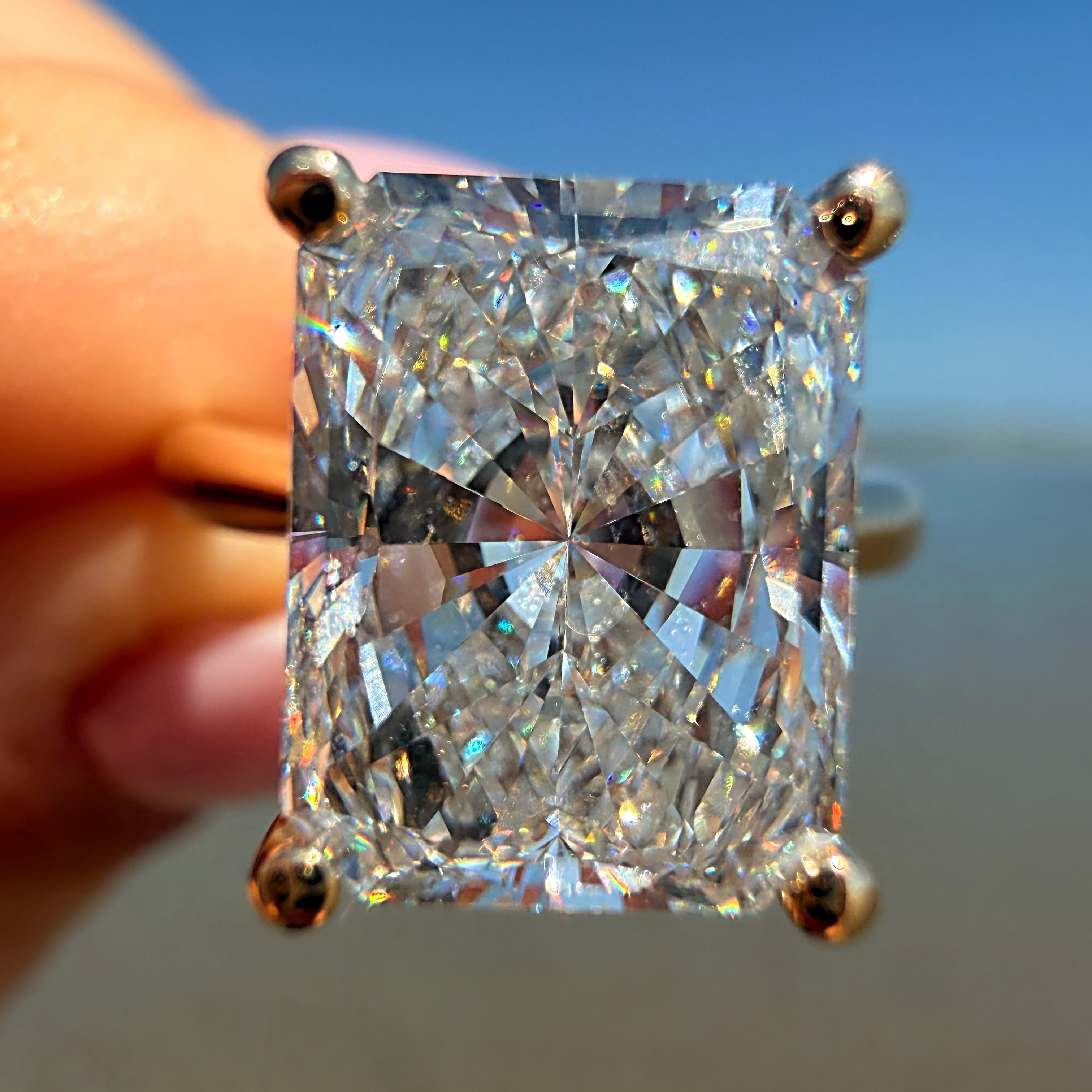 A radiant cut diamond ring held by a hand against a blurred beach and blue sky background.