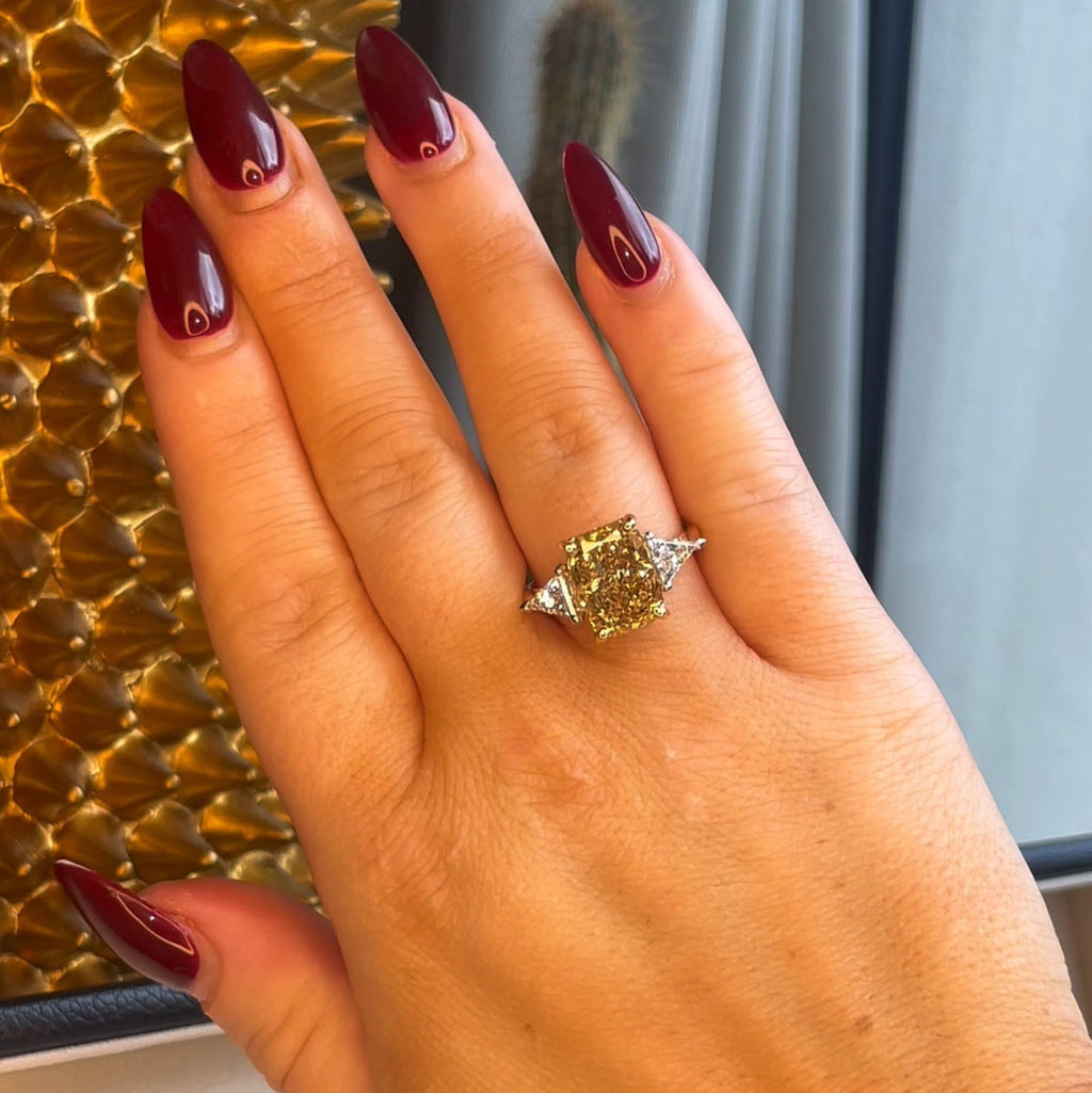 Hand wearing a yellow gemstone ring with red nail polish against a textured wall.