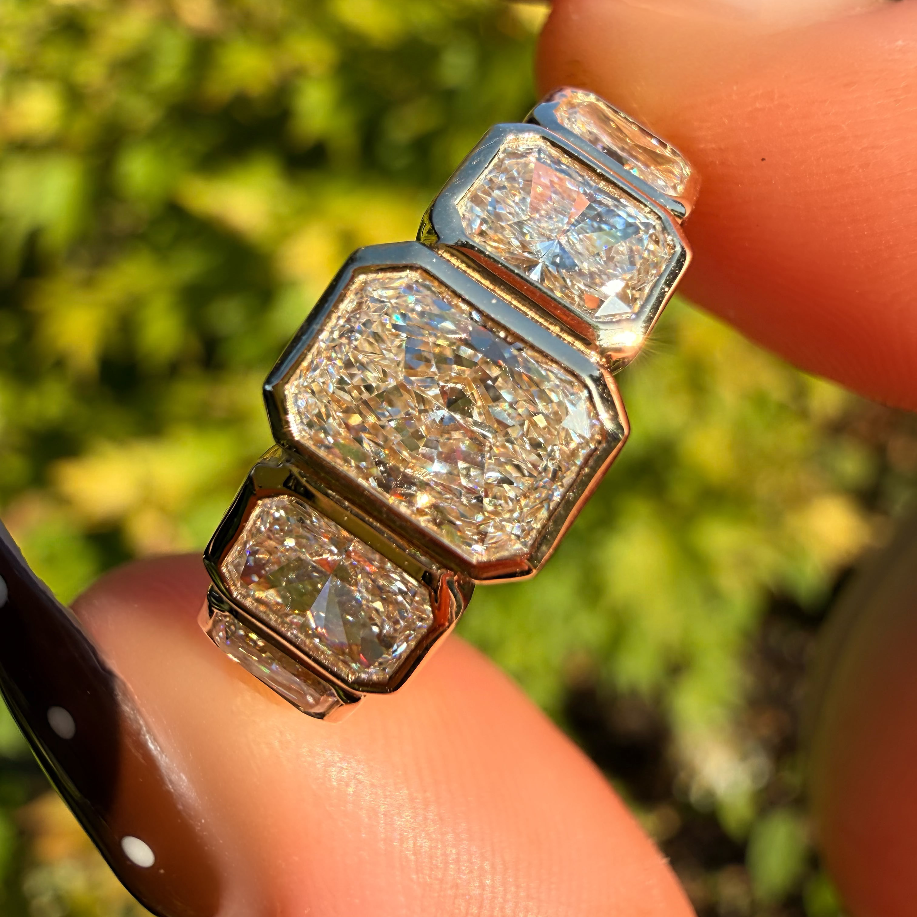 A close-up of a silver-toned ring with three stacked rectangular clear gemstones, held between fingers against a blurred green outdoor background.