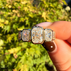 A close-up of a hand holding a vintage-style ring with three rectangular baguette-cut clear stones in a gold-toned setting, with a blurred green and yellow outdoor background.